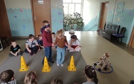 dzieci siedzą na dywanie na środku stoi chłopiec z opaską na oczach naywanie stoją trzy pachołki przed dywanem leży obręcz a w niej pluszowa żyrafa