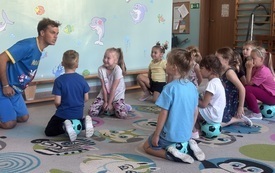 grupa dzieci klęczy na dywanie dzieci się śmieją przed nimi klęczy pan w śmiesznej pozie w niebieskim stroju sportowym drużyna kangura