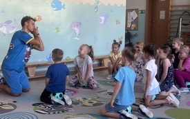 grupa dzieci klęczy na dywanie przed nimi klęczy pan w śmiesznej pozie w niebieskim stroju sportowym drużyna kangura
