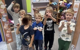 Sześcioro dzieci stoi w sali z p&oacute;łkami pełnymi zabawek, trzymając w dłoniach małe przedmioty.