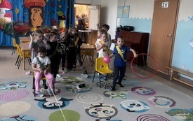 Dzieci bawią się w sali przedszkolnej, trzymając obręcze i pałeczki, a na dywanie leżą kolorowe piłki i obręcze.