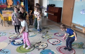 Dzieci bawią się w sali przedszkolnej na kolorowym dywanie, używając lasek z czerwono-białymi paskami; w tle widoczne są pianino i dekoracja jesienna
