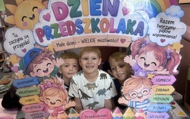Trzech chłopc&oacute;w uśmiecha się do zdjęcia przez kolorową ramkę z napisem Dzień Przedszkolaka oraz ilustracjami dzieci, słońca, tęczy i kwiat&oacute;w.