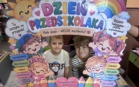 Dw&oacute;ch chłopc&oacute;w pozuje za kolorową tablicą z napisem Dzień Przedszkolaka, ozdobioną tęczą, słońcem, sercami i postaciami dzieci.