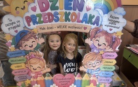 Dwie dziewczynki pozują za kolorową planszą z napisem Dzień Przedszkolaka, ozdobioną ilustracjami dzieci, tęczą, kwiatami i hasłami o przedszkolu.