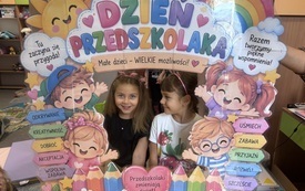 Dwie dziewczynki pozują za kolorową planszą z napisem Dzień Przedszkolaka i ilustracjami dzieci, słońca, tęczy oraz kwiat&oacute;w.