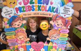 Dwoje dzieci pozuje za kolorową dekoracją z napisem Dzień Przedszkolaka, przedstawiającą uśmiechnięte postacie, kwiaty i hasła o przyjaźni, zabawie oraz rozwoju.