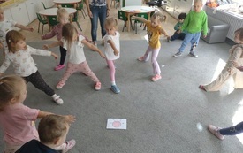 grupa dzieci stoi na dywanie i wykonuje zadanie ruchowe z obrazka kt&oacute;ry leży na środku