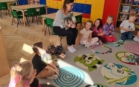 dzieci siedzą w kole a wśr&oacute;d nich pani siedzi na krzesełku z otwartą książką i pokazuje w niej coś dzieciom