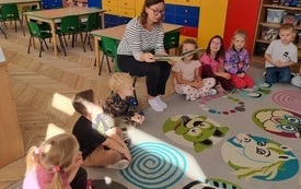 dzieci siedzą w kole a wśr&oacute;d nich pani siedzi na krzesełku z otwartą książką