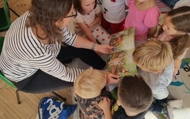 dzieci stłoczone na dywanie przed kobietą kt&oacute;ra trzyma otworzoną książkę