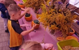 dzieci stoją wzdł&oacute;ż długiego stołu przed nimi stoją małe wydrążone dynie z gąbką florystyczną w środku oraz jesienne kwiaty