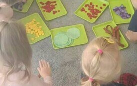 dzieci siedzą na dywanie przed nimi ustawione są tacki z r&oacute;żnej wielkości i kolorystyce k&oacute;łkami