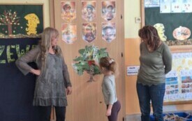 dzieci siedzą na dywanie dwie nauczycielki stoją jedna dziewczynka przypina sylwetę jabłka do szablonu drzewa zawieszonego na drzwiach