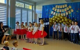 trzy dziewczynki z mikrofonami na środku sali w tle pozostałe dzieci po bokach panie nauczycielki z gitarami 