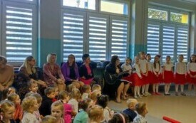 dzieci i panie nauczycielki obserwują występ widać panią z gitarą i dziewczynki w odświętnych strojach