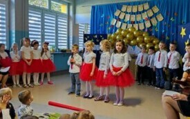 trzy dziewczynki i chłopiec z mikrofonami na środku sali w tle pozostałe dzieci po bokach panie nauczycielki z gitarami 