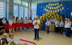 chłopiec w odświętnym stroju na środku sali trzyma mikrofon w tle pozostałe dzieci 