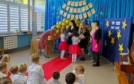 dwie dziewczynki z grupy V wręczają wyr&oacute;żnienia swoim paniom nauczycielkom obok pani dyrektor i inne nauczycielki obserwujące dzieci z sali