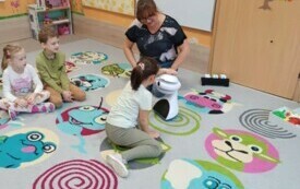 dzieci siedzą na dywnie dziewczynka dotyka robota za nimi nauczycielka