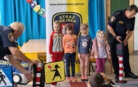dzieci stoją przed przejściem dla pieszych chłopiec przebrany za pojazd straży miejskiej stoi przed pasami strażnicy miejscy po bokach