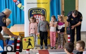 dzieci stoją przed przejściem dla pieszych chłopiec przebrany za pojazd straży miejskiej stoi przed pasami strażnicy miejscy po bokach