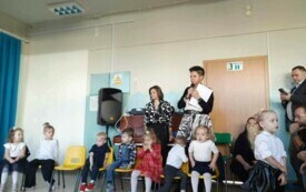 grupka dzieci, za nimi pani dyrektor z mikrofonem, widać rodzic&oacute;w