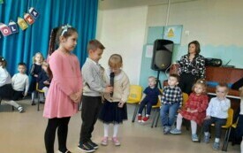 troje dzieci stoi na środku sali, jedno z nich z mikrofonem, w tle dekoracja pasowanie na przedszkolaka i inne dzieci siedzące na krzesełkach