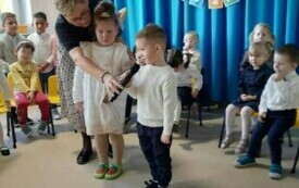 dwoje dzieci stoi na środku sali, nauczycielka stoi obok z mikrofonem, w tle dekoracja pasowanie na przedszkolaka i inne dzieci siedzące na krzesełkach