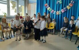 grupka dzieci stoi na środku sali, jedno z nich z mikrofonem, w tle dekoracja pasowanie na przedszkolaka i inne dzieci siedzące na krzesełkach