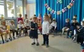 dwoje dzieci stoi na środku sali, jedno z nich z mikrofonem, w tle dekoracja pasowanie na przedszkolaka i inne dzieci siedzące na krzesełkach