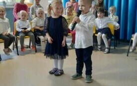 dwoje dzieci stoi na środku sali, jedno z nich z mikrofonem w tle dekoracja pasowanie na przedszkolaka i inne dzieci siedzące na krzesełkach