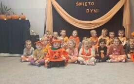 grupa dzieci w pomarańczowych strojach z wymalowanymi buziami obok pani nauczycielka w pomarańczowym przebraniu na tle dekoracji święto dyni