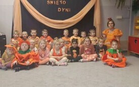 grupa dzieci w pomarańczowych strojach z wymalowanymi buziami obok pani nauczycielka w pomarańczowym przebraniu na tle dekoracji święto dyni