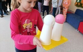dziewczynka niesie tacę na kt&oacute;rej stoją dwa kubki a na nich leżą piłeczki w tle dzieci