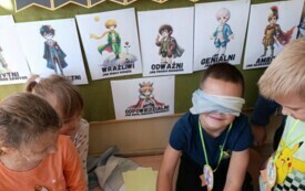 chłopiec z przewiązanymi biała chustką oczami rysuje coś na kartce, dzieci przygądają się w tle napis dzień chłopaka i postacie na kartkach