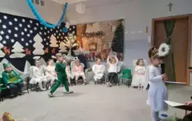 dziewczynka w przebraniu aniołka z mikrofonem na pierwszym planie  nieco z tyłu chłopiec w zielonym przebraniu inne dzieci w tle siedzą na krzesełkach dookoła świąteczna dekoracja