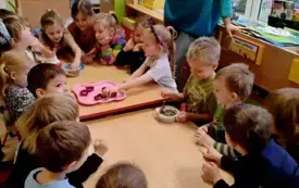 grupa dzieci siedzi przy złączonych stołach chłopiec i dziewczynka trzymają miseczki z ziarnem na stole taca z foremkami