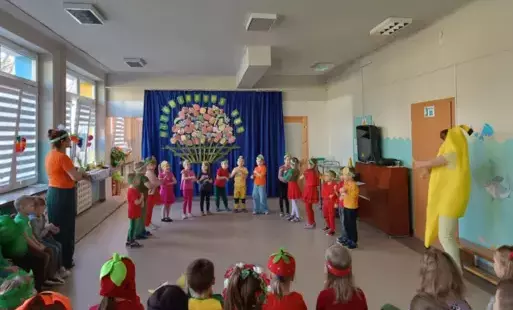 dzieci w strojach owocowo warzywnych z opaskami na głowach stoją w półkolu po bokach nauczycielki w przebraniach w tle dekoracja kosz z owocami i warzywami 