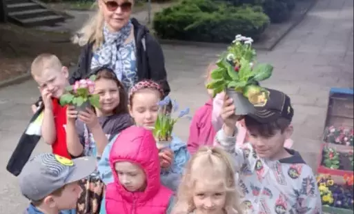 grupa dzieci stoi na chodniku wraz z panią nauczycielką obok kolorowy pociąg wykonany ze skrzynek a w skrzynkach kolorowe kwiaty