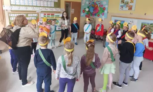dzieci stoją w kole z nimi dwie panie nauczycielki jedna z nich w ubiorze pszczelarza dzieci mają na głowach żółte opaski z pszczółkami