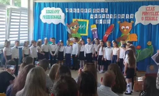 dzieci w odświętnych strojach stoją obok siebie, w tle dekoracja żegnamy przedszkole, czas pomyśleć o szkole