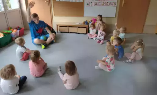 dzieci siedzą na dywanie w kole wśród nich pan ubrany na niebiesko z uniesioną w górzę ręką
