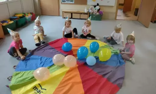 dzieci siedzą przed chustą animacyjną leżącą na podłodze na chuście leżą kolorowe balony