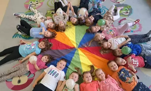 dzieci leżą w kole na chuście animacyjnej głowami do środka niektóre z dzieci trzymają balony