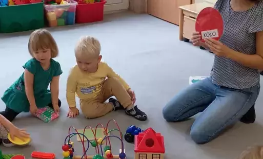 dzieci siedzą na dywanie przed nimi pomoce dydaktyczne w kształcie kółek pani pokazuje dzieciom czerwone koło z napisem koło