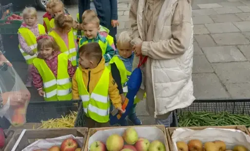 grupa dzieci w kamizelkach odblaskowych stoi wraz z paniami przed skrzynkami wypełnionymi jabłkami, fasolką i pomidorami