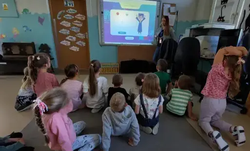 dzieci siedzą na dywanie przed nimi tablica multimedialna z postacią animowaną pani stoi obok tablicy twarzą zwróconą do dzieci