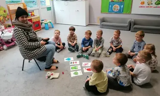 dzieci siedzą w półkolu przed nimi leżą na dywanie obrazki z elementami garderoby pani siedzi na krzesełku w ubraniu zimowym