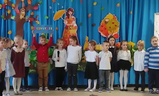 dzieci w eleganckich strojach stoją w półkolu chłopiec trzyma w górze tabliczkę z napisem wszystkie w tle dekoracja jesienna